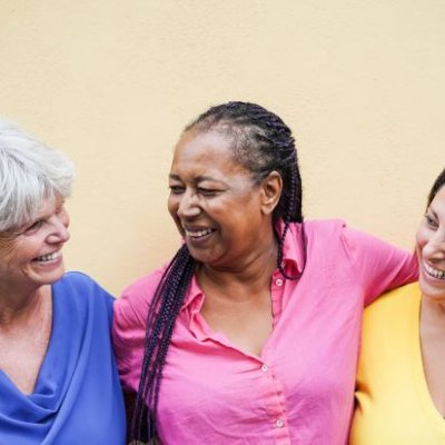 Three women in brightly coloured shirts smile and laugh. They have their arms around each other.